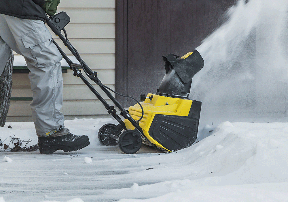 A closeup of a man in winter gear using an electric snowblower to clear a snow-covered driveway.