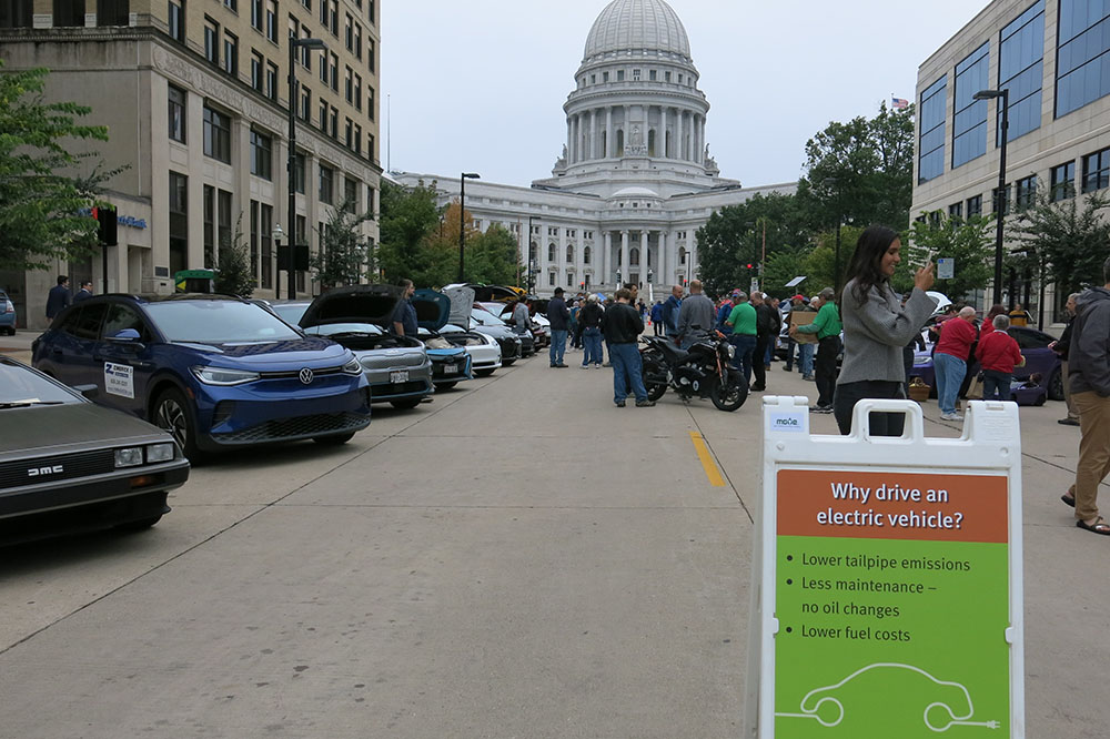 A group of people congregate around a row of electric vehicles for Madison's Drive Electric Week event with the Capitol building in the background.