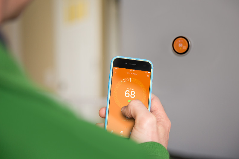 A closeup of a cell phone on a smart thermostat app with an orange background at 68 degrees, with an orange thermostat on the wall in the background.