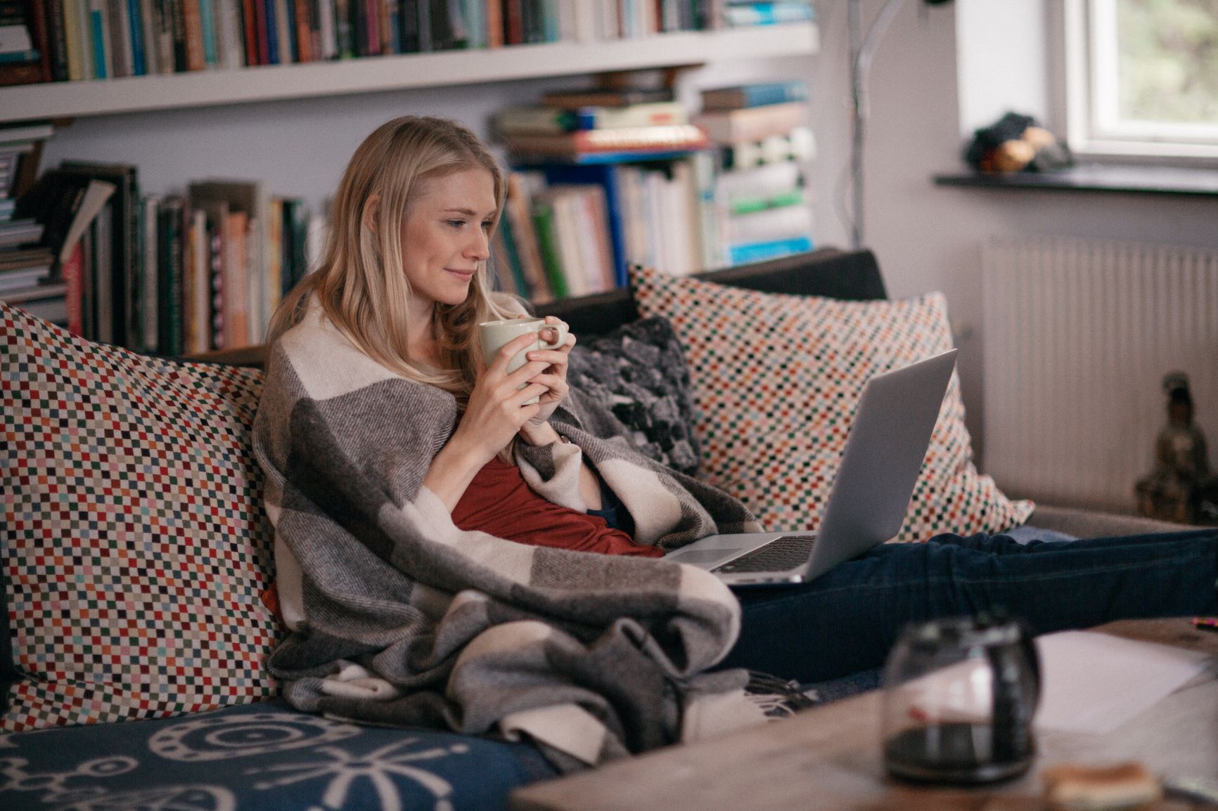 woman on a couch with blanket and coffee looking at laptop