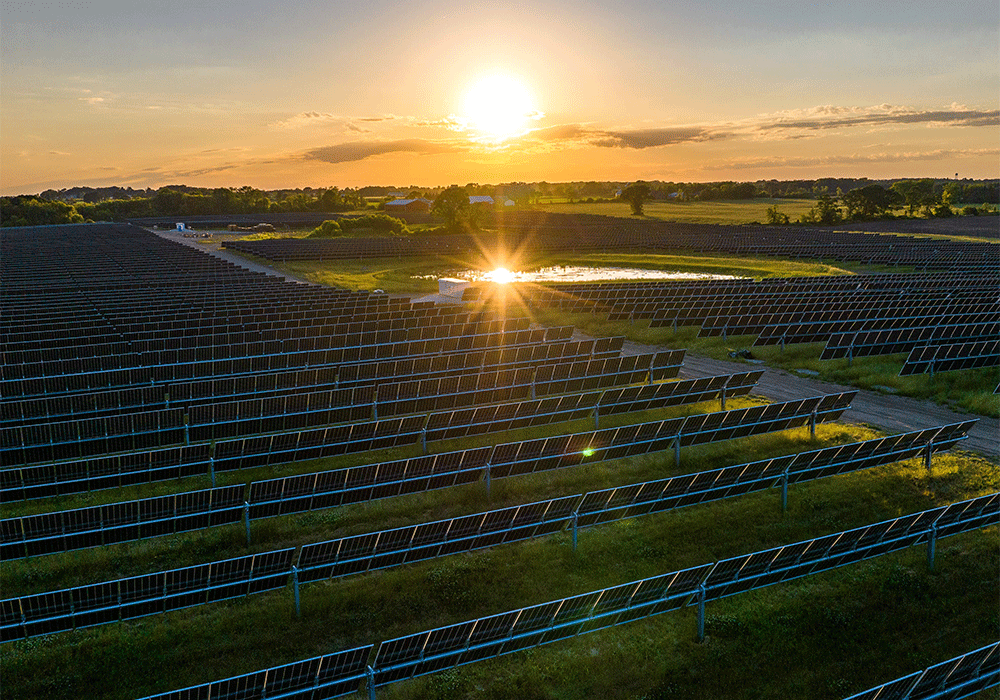 A drone's view of the solar array at Paris Solar with green grass and fields in the background and a bright sun along the horizon.